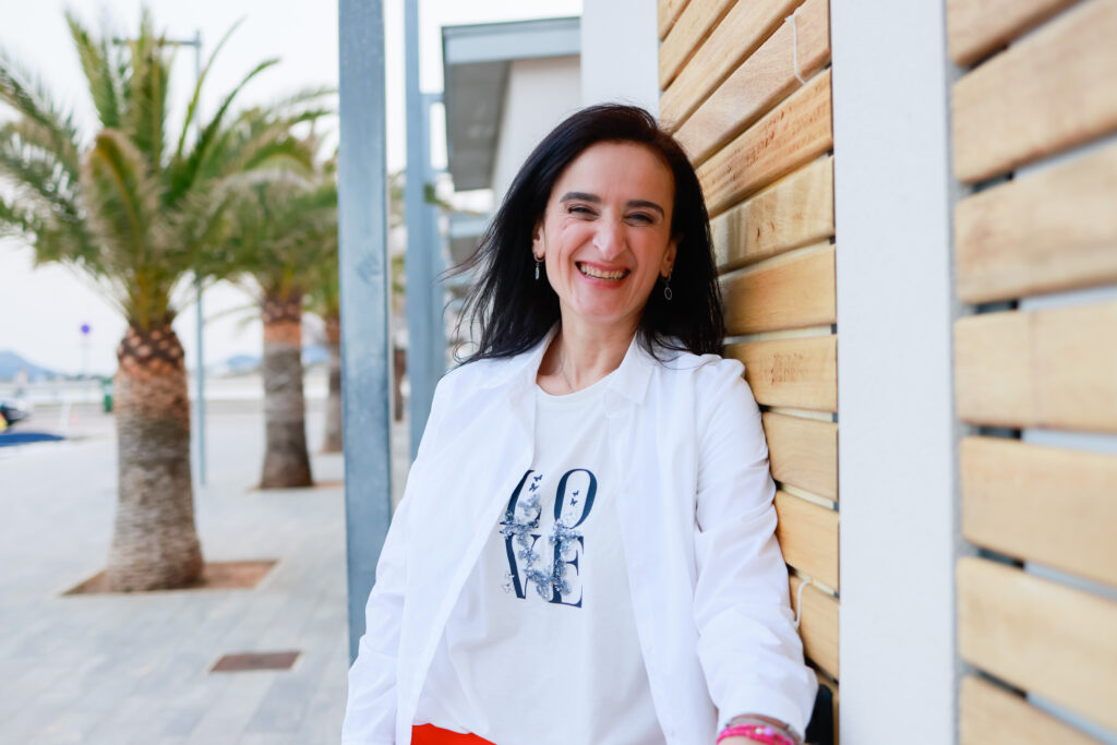 Sevtap Züberi trägt einen weißen Blaser, steht vor einer Holzwand und lächelt direkt in die Kamera.