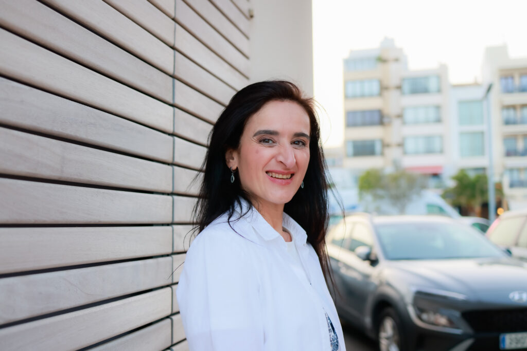 Sevtap Züberi trägt einen weißen Blaser, steht vor einer Holzwand und lächelt seitlich in die Kamera.