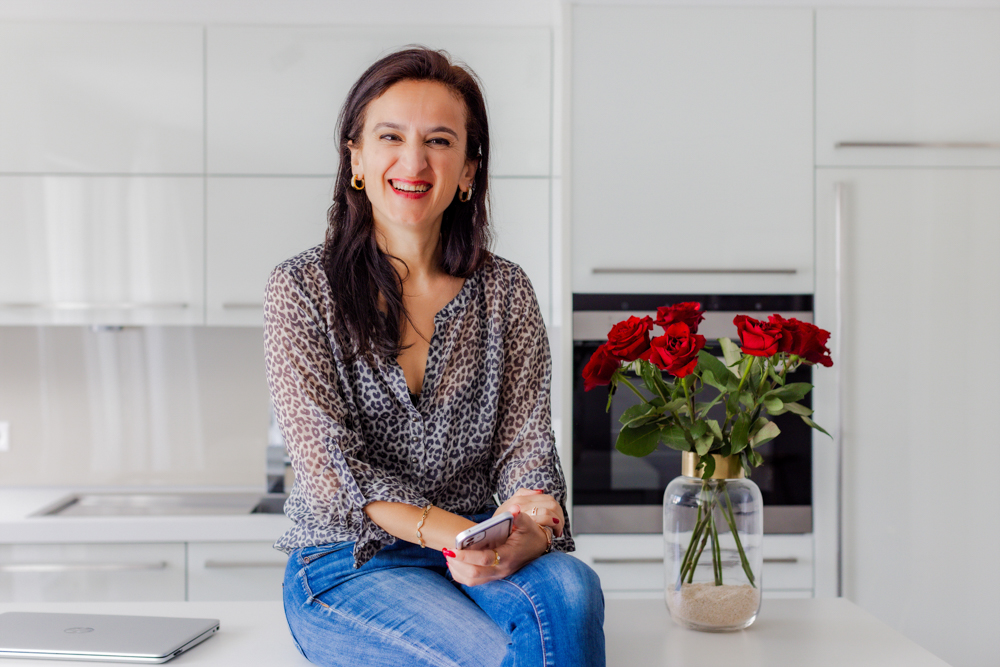 Sevtap Züberi sitzt lässig in Jeans und schicker Bluse auf einem Küchentresen und hat die Beine übereinandergeschlagen. Neben ihr ist ein Strauß roter Rosen zu sehen. Sie hält ein Handy in der Hand.