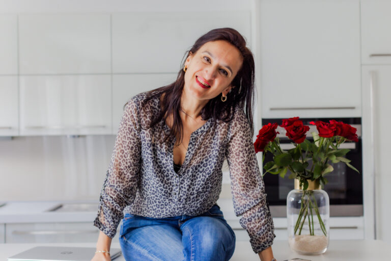 Sevtap Züberi sitzt lässig in Jeans und schicker Bluse auf einem Küchentresen und hat die Beine übereinandergeschlagen. Neben ihr ist ein Strauß roter Rosen zu sehen. Sie neigt den Kopf und strahlt.
