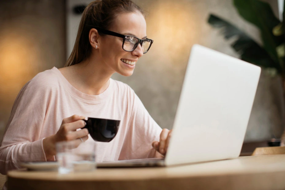 Eine Frau sitzt am Laptop mit einer Kaffeetasse in der Hand und sieht fröhlich aus.