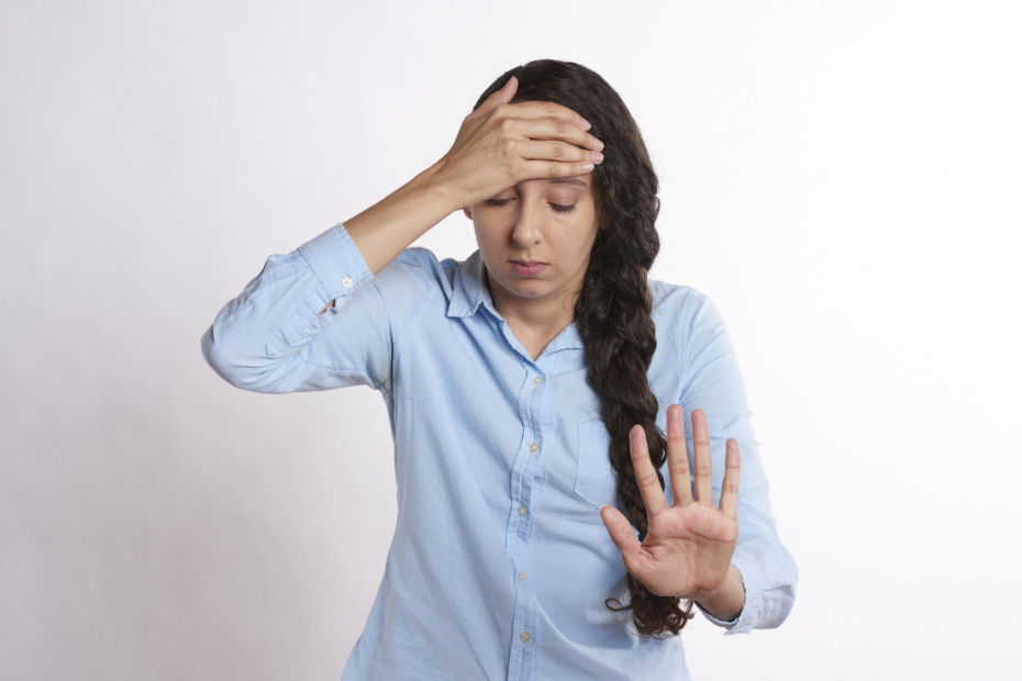 Frau hält sich mit einer Hand die Stirn und mit der anderen wehrt sie ab. Ihr Blick ist nach unten gerichtet.